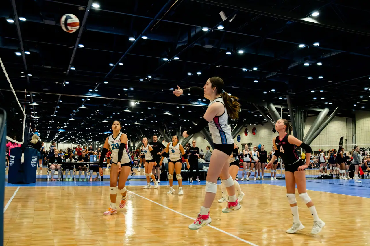 DS Elite server in a white uniform in mid-air on her follow-through as teammates watch, under a ceiling full of lights