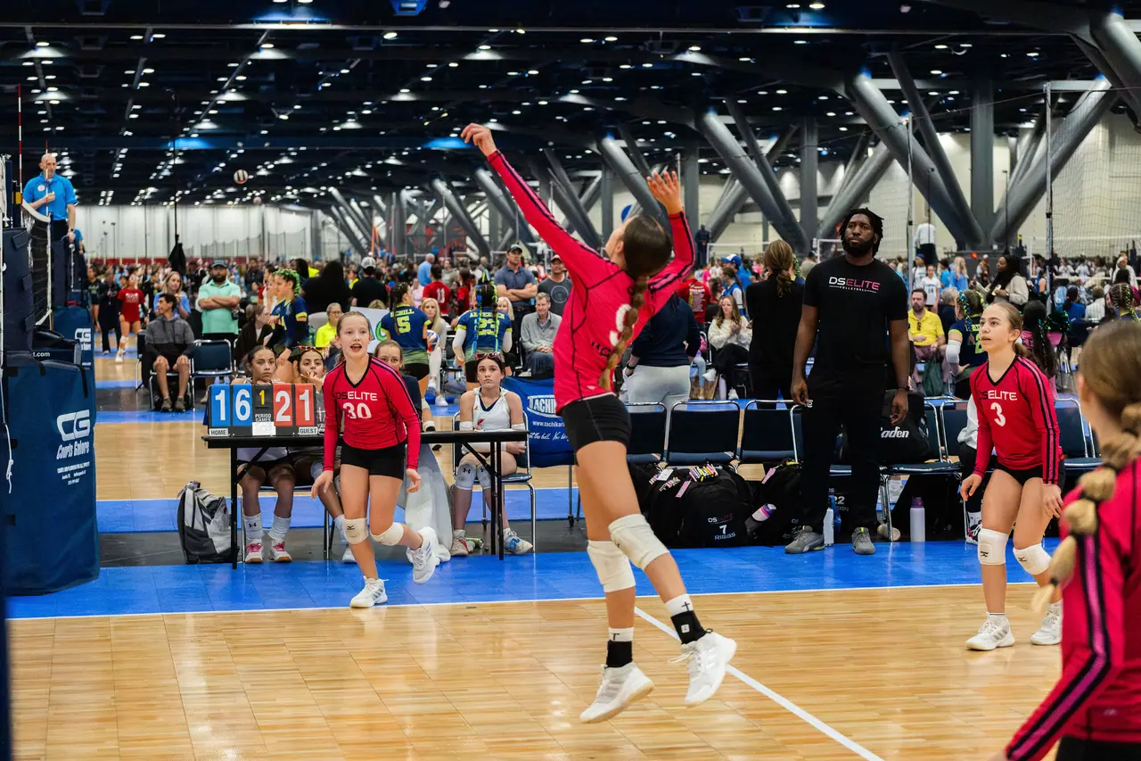 Wide view of a DS Elite server in the air at the end line, with the scoreboard, a sideline coach and a huge tournament hall behind