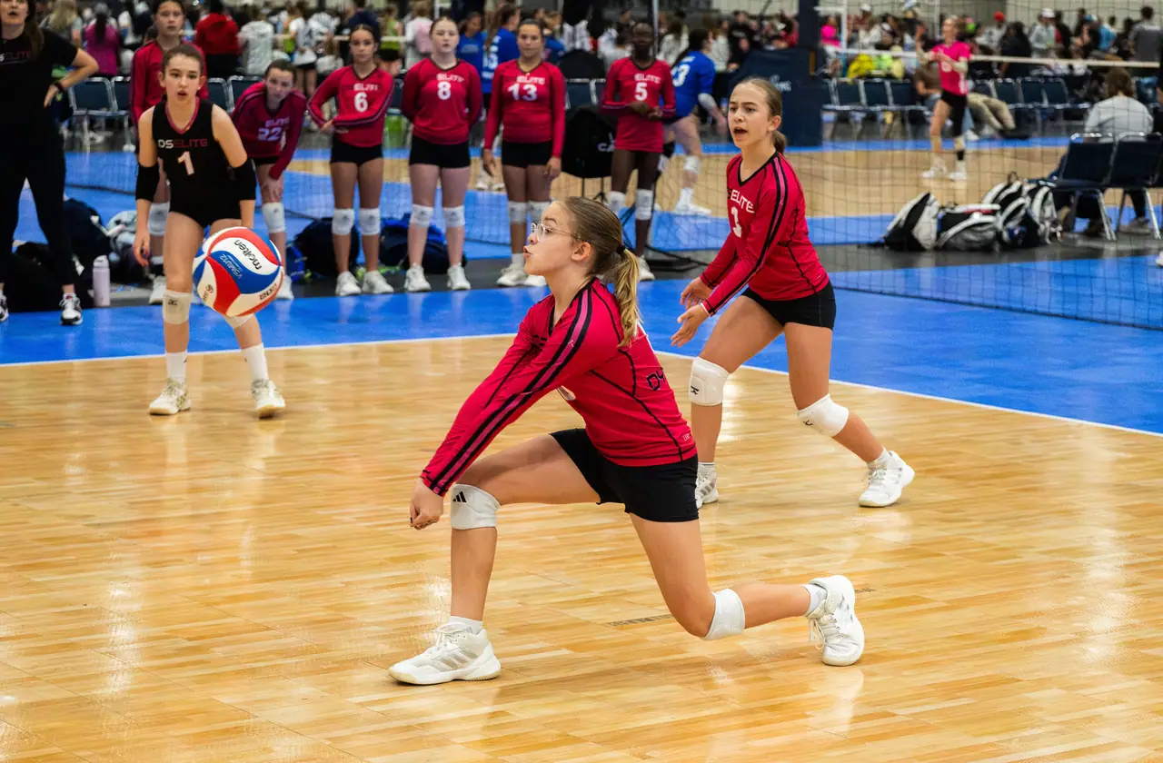 DS Elite player in a raspberry uniform lunging low to pass the ball, with her bench behind her