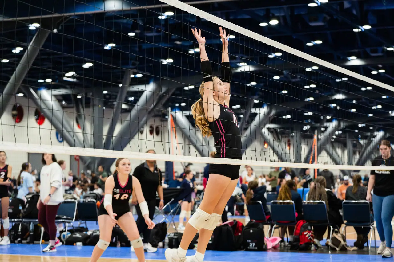 DS Elite player in a black uniform leaping at the net with both hands up to block, seen through the net