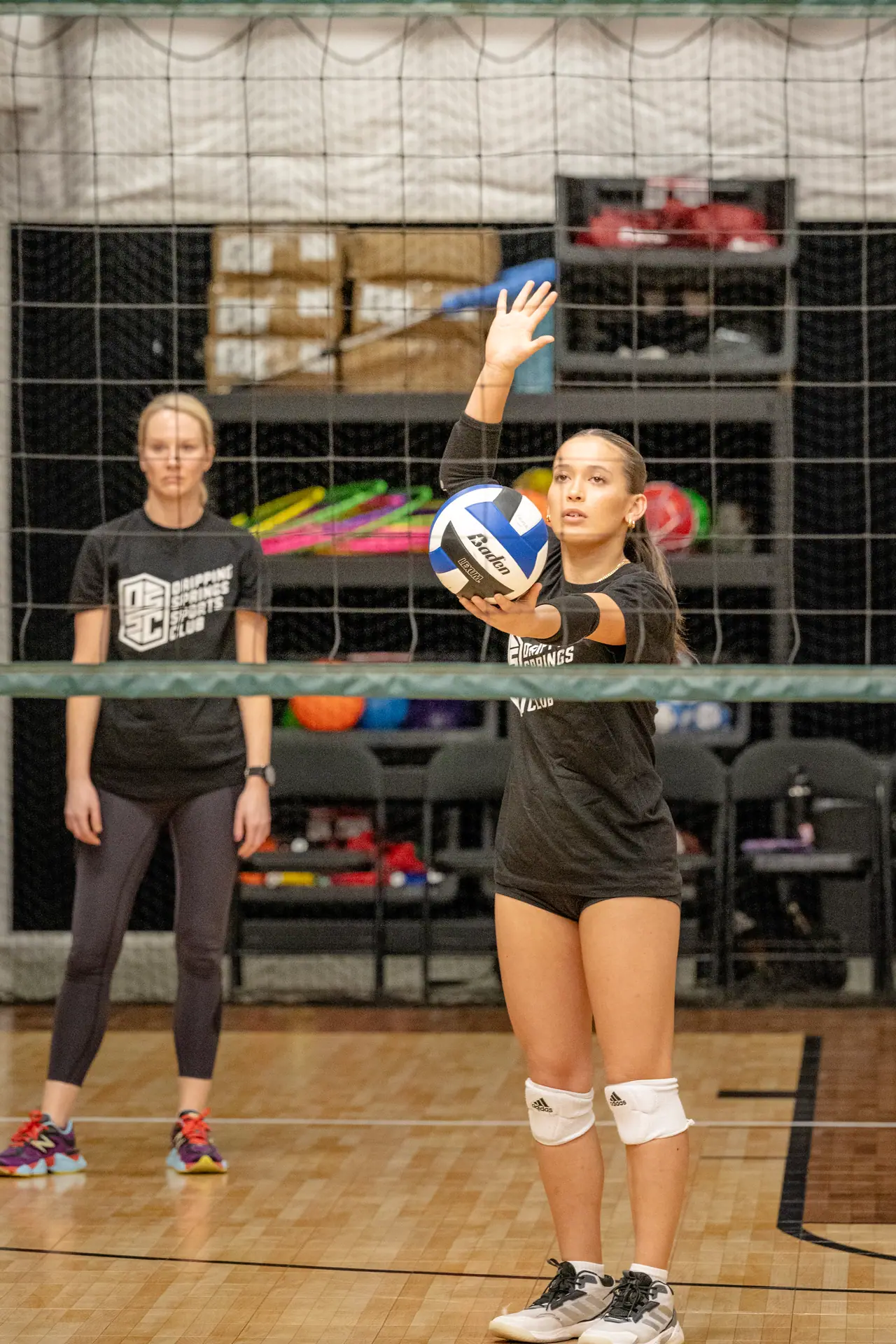 Player tossing the ball to serve during a training session as a coach watches, shot through the net