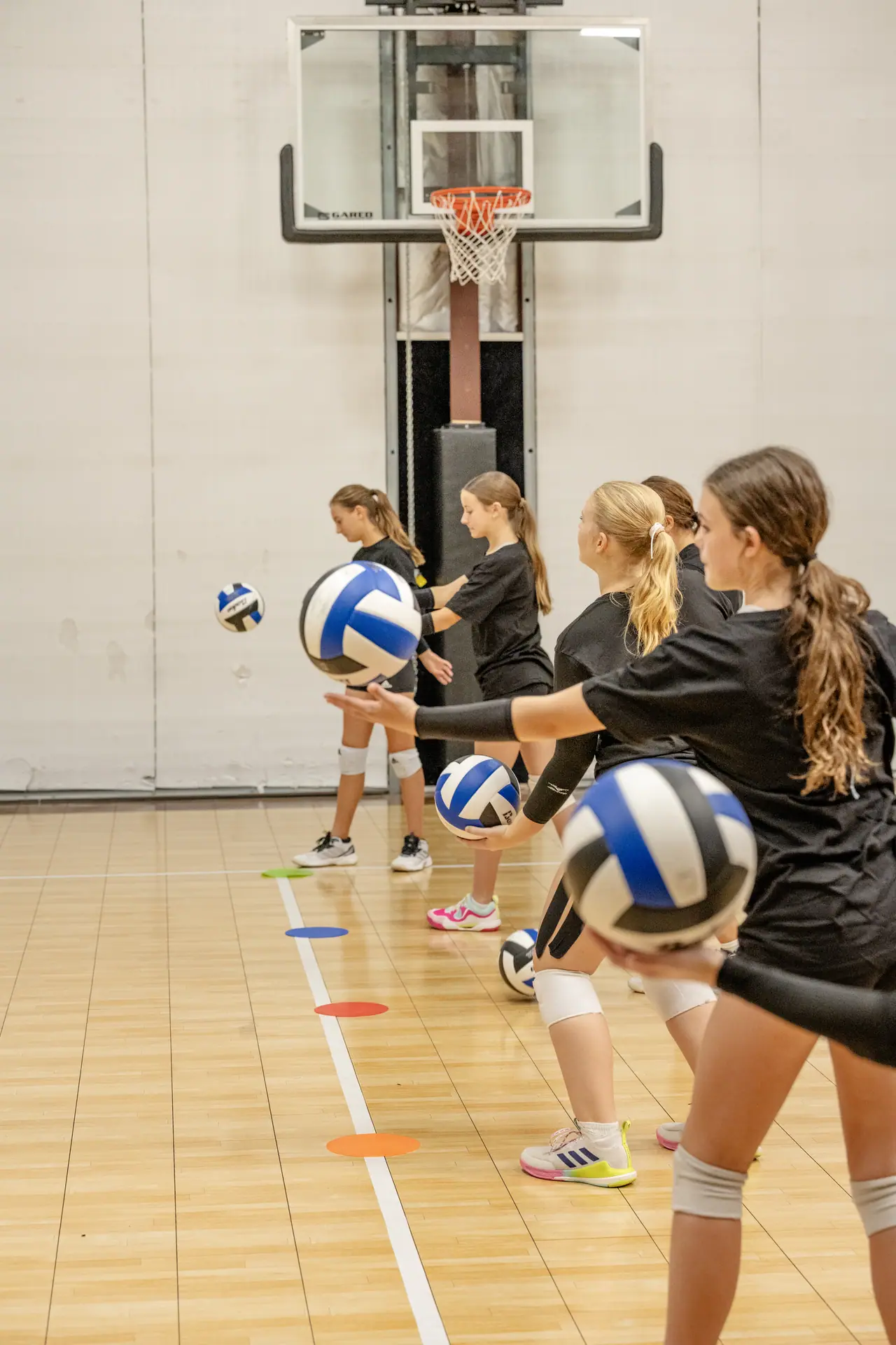 Players in black practice shirts lined up on the court, each tossing a volleyball in a serving drill at the DSSC gym